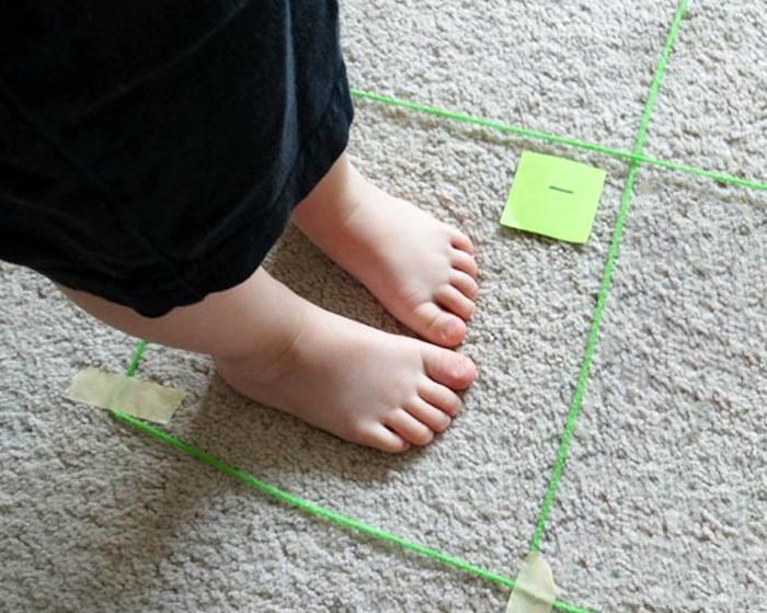 Image shows a kid standing in a square made with yarn on top of the carpet. From KAB.